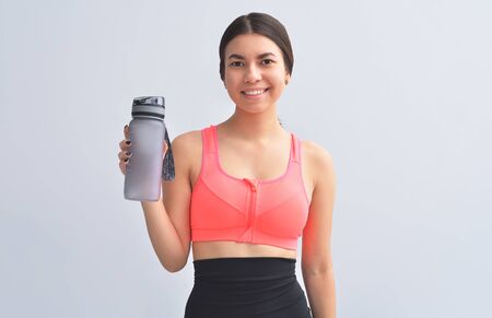 Restoring water balance. Attractive and young woman in sports wear keeping bottle in hand and smiling while standing on gray backgroundの写真素材