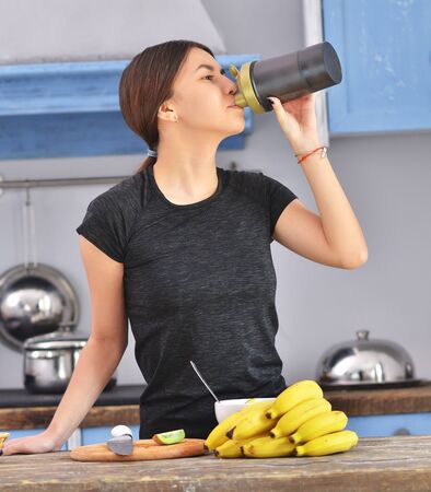 Young woman drinking protein shake near table with ingredients in kitchenの写真素材