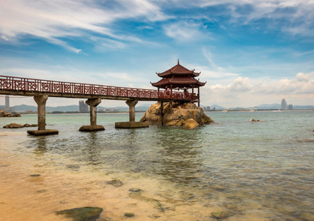 Traditional Asian-style pavilion perched on a rock and connected to the beach by a red ornate bridge, surrounded by calm sea waters and distant skylineの写真素材