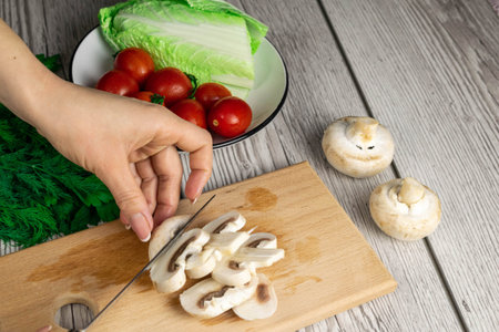 Female hand cuts mushrooms on a wooden board on a wooden table with herbs and cherry tomatoesの写真素材