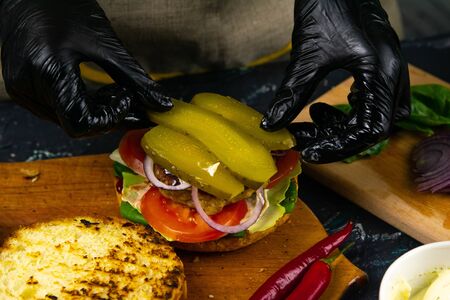 Process of cooking burgers. Cropeed view of chef hands in black gloves with prepared cheeseburger, variety of fillings and ingridients on wooden desk. Catering and Good Quality Fastfood conceptの写真素材
