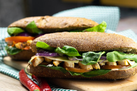 A dark bread sandwich with chicken, tomatoes, spinach, cheese and carrots stands on a wooden board...