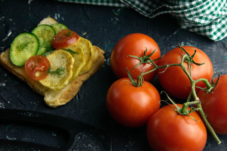 Sandwich with potatoes, cucumber and tomato next to tomatoes on a branch, stand on a black table....