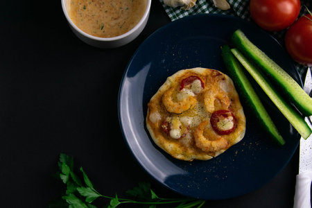 Small mini pizza with shrimp, tomato and cheese on blue plate with cucumber around a plate of...