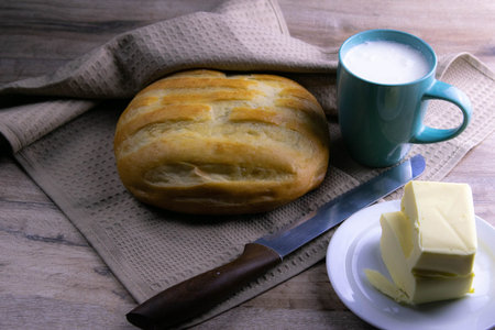 A loaf of bread under a towel on the table with a glass of milk and butter. Top views