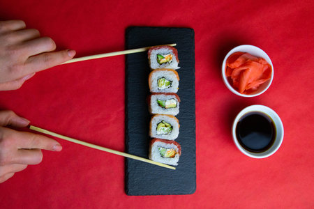 Red and orange rolls on a granite board for sushi, next to Women's hands hold all the rolls with sticks, ginger and soy sauce, all standing on a red background. Top viewsの写真素材