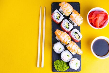 Granite board for sushi, filled with rolls of different kinds, next to stand sticks for sushi, red marinaginger and soy sauce on a orange background. Top views with clear spaceの写真素材