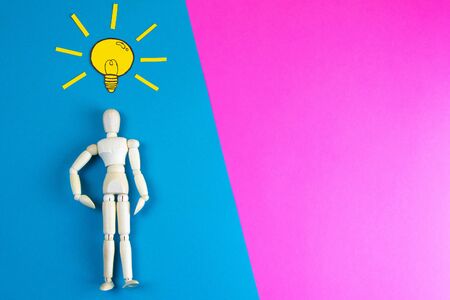 Wooden figurine of a man with a light bulb with yellow craft paper over his head on a blue and pink background with clear space for text.の写真素材