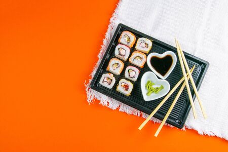 Black plate with sushi, filled with rolls of different kinds, next to stand sticks for sushi, red marinaginger and soy sauce on a orange background. Top views withの写真素材