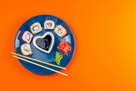 Blue plate with sushi, filled with rolls of different kinds, next to stand sticks for sushi, red marinaginger and soy sauce. on a orange background. Top viewsの写真素材