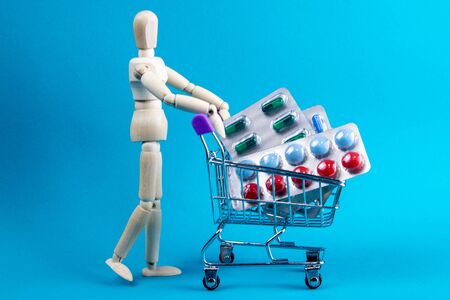 Wooden doll and metal shopping cart with pills on blue background. Various capsules, tablets and medicine on blue pastel trendy background. Pills concept.の写真素材