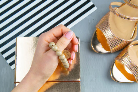 A woman's hand holds a bracelet on her finger over a gold notepad, next to them are gold shoes.