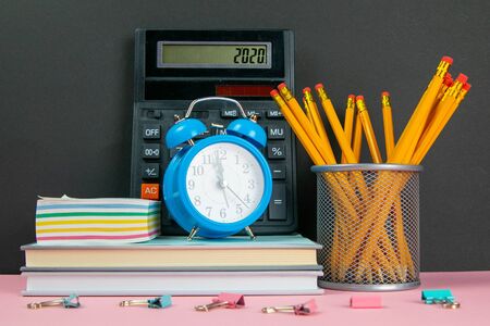 On a stack of books there is a calculator, a clock and sheets for notes, and next to a glass of pencils.の写真素材