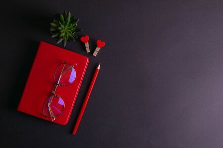 The red notebook stands on a black background, on the notebook there are glasses and next to the notebook, a flower, two clothespins in the shape of hearts and a red pencil.の写真素材