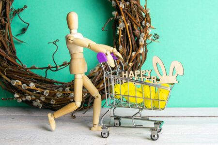 The Easter concept. Figure of a wooden man with a basket filled with eggs. In the background is a wooden rabbit and a sign of a Happy Easter and a wreath.の写真素材