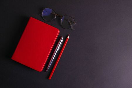 The red notebook stands on a black background, next to the notebook are glasses, a red pencil and a silver pen.の写真素材