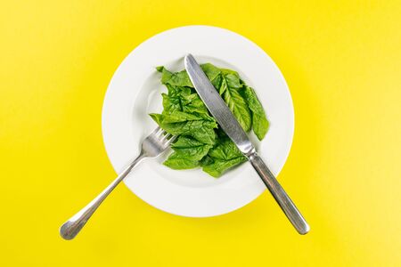 Fresh green spinach leaves on a white plate fork knife on bright yellow background flat lay top view copy space. Baby young spinach leaves, healthy food, diet. Creative food concept.の写真素材