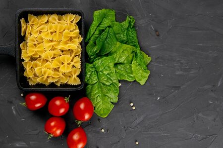 Raw pasta farfalle in frying pan, tomatoes and spinach on dark background. Top views with clear space.の写真素材