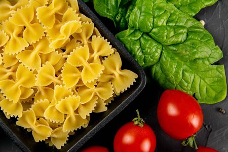 Raw pasta farfalle in frying pan, tomatoes and spinach on dark background. Top views, close-up.の写真素材
