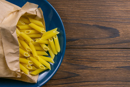 Pene Lisce pasta in a craft paper bag on a blue plate on a wooden table. Top views with clear space.