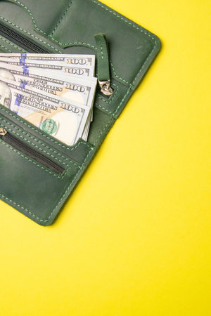 Several hundred dollar bills protrude from a stylish green leather wallet on a bright yellow...