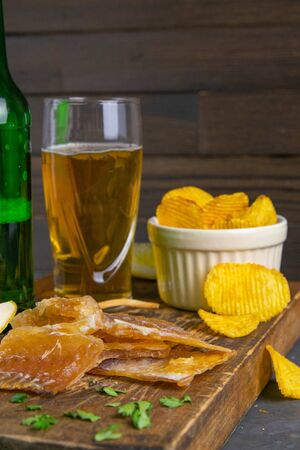 Dried cod fillet with beer, lemon and potato chips on dark wooden board. Snack on fish with beer. Front views, close-up.の写真素材