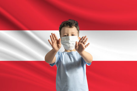 Little white boy in a protective mask on the background of the flag of Austria. Makes a stop sign with his hands, stay at home Austria.の写真素材