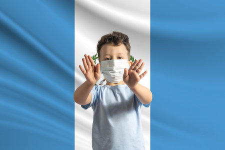 Little white boy in a protective mask on the background of the flag of Guatemala. Makes a stop sign with his hands, stay at home Guatemala.の写真素材