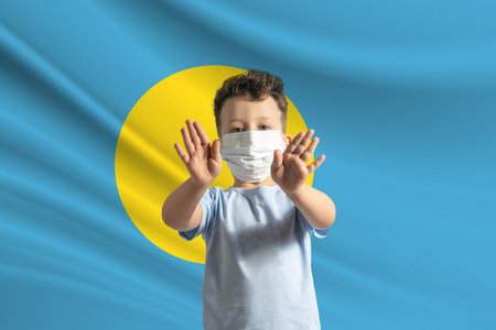 Little white boy in a protective mask on the background of the flag of Palau. Makes a stop sign with his hands, stay at home Palau.の写真素材