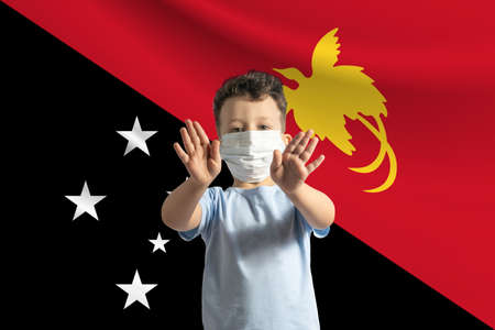 Little white boy in a protective mask on the background of the flag of Papua New Guinea. Makes a stop sign with his hands, stay at home Papua New Guinea.の写真素材