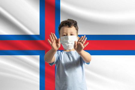 Little white boy in a protective mask on the background of the flag of Faroe Islands. Makes a stop sign with his hands, stay at home Faroe Islands.の写真素材