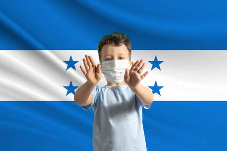 Little white boy in a protective mask on the background of the flag of Honduras. Makes a stop sign with his hands, stay at home Honduras.の写真素材
