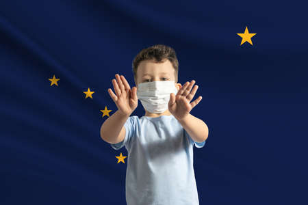 Little white boy in a protective mask on the background of the flag of Alaska. Makes a stop sign with his hands, stay at home Alaska.の写真素材