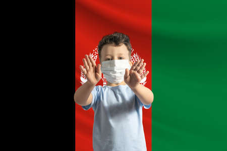 Little white boy in a protective mask on the background of the flag of Afghanistan. Makes a stop sign with his hands, stay at home Afghanistan.の写真素材