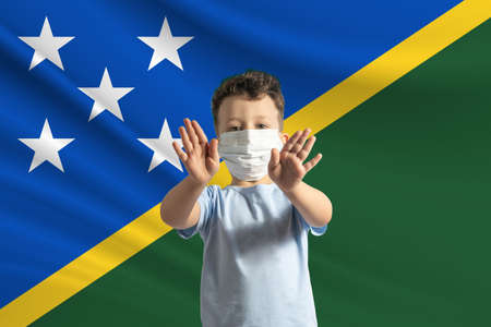 Little white boy in a protective mask on the background of the flag of Solomon Islands. Makes a stop sign with his hands, stay at home Solomon Islands.の写真素材