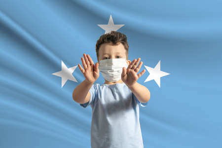 Little white boy in a protective mask on the background of the flag of Federated States of Micronesia. Makes a stop sign with his hands, stay at home Federated States of Micronesia.の写真素材