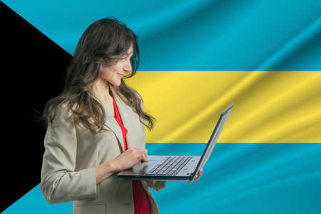 Freelance in Bahamas. Beautiful young woman freelancer uses laptop computer against the background of the flag of Bahamas.の写真素材