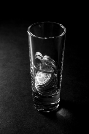 A glass for alcoholic beverages filled with Euro coins. The glass stands on a dark table with a black background. alcoholism.の写真素材