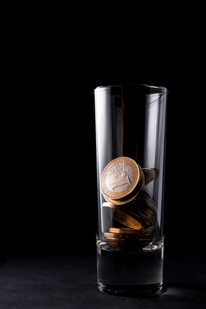 A glass for alcoholic beverages filled with Euro coins. The glass stands on a dark table with a black background. alcoholism.の写真素材
