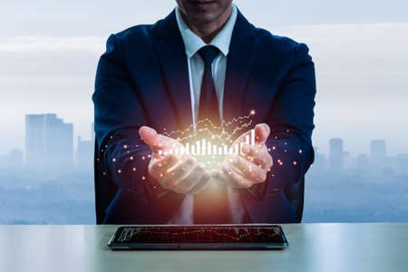 In hand of a man wearing a suit There is a stock chart that is doing new heights. On a tabletop with tablet on the screen, there are green and red stock graphs, the backdrop of the cityscape blurred.の写真素材