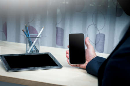A man wearing a suit and sitting on a chair Use your right hand to hold a black screen mobile phone and a black screen tablet placed on the table.の写真素材