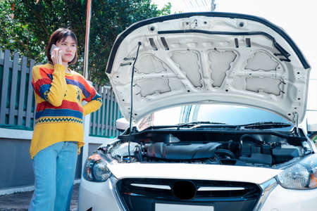 A woman standing next to a car with a bonnet opened and using a cell phone to contact a mechanic to fix the damaged car.の写真素材