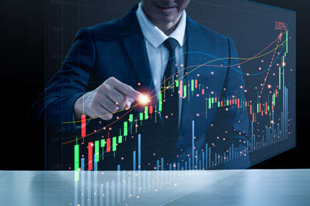 An Asian man in a suit is sitting at a desk with his right hand pointing at a hologram showing a graph of stocks going up to the top. Black background blurの写真素材