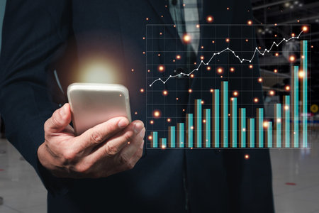 A man in a suit holds a mobile phone with his right hand The screen lights orange. The side area has a stock graph that is peaking and the background is blurred.の写真素材
