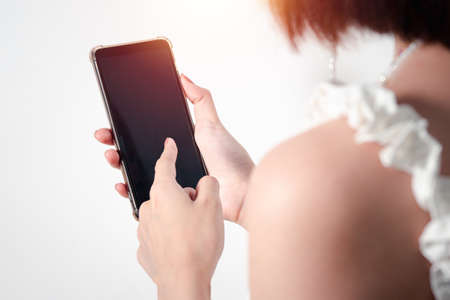 A young Asian woman uses her right hand to hold a mobile phone with a black screen (focus) and her left hand (blur) pointing at the screen. White background blurの写真素材