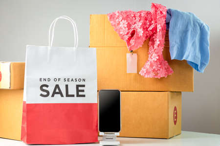 A mobile phone with a black screen was placed next to a white-red shopping paper bag. Behind the scenes clothes were placed in a blurred box.の写真素材