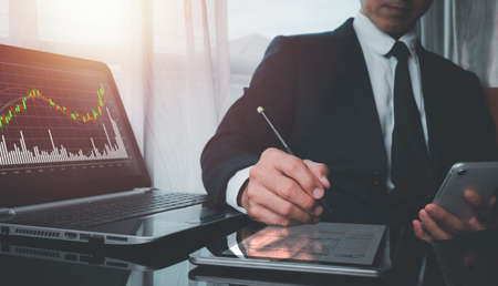 A man wearing a suit on his left hand holding a cell phone His right hand poked a pen on a tablet's screen, and the screen of the laptop shows the fast-growing stock chart.の写真素材
