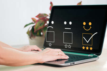 On a laptop computer, a woman selects a facial smiling emoticon to display on the virtual screen. The user experience or customer satisfaction survey, poll, or questionnaire.の写真素材