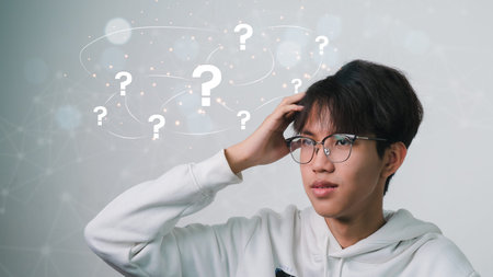 Question mark above the head of a contemplative Asian man wearing glasses on a gray backdrop.no...