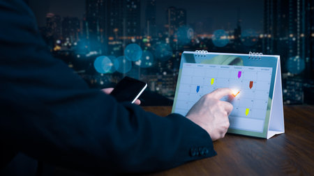 Timetable, agenda, and schedule for the 2025Â event planner. A businessman at an office desk takes notes on the calendar while using his phone to check the planner. Using calendars and event organizingの写真素材